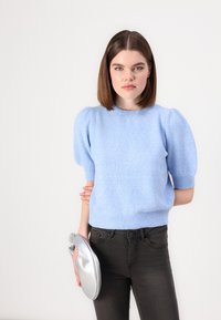 Maglione lavorato a maglia azzurro chiaro con maniche a palloncino e orlo a coste, abbinato a jeans scuri. Tiene in mano una borsa a tracolla argentata e lucida.