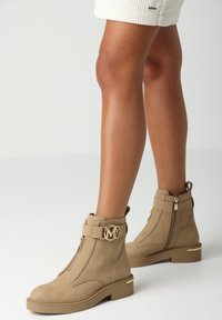 Kahové kotníkové boty vyrobené z hladké kůže, s zlatým logo detailem, bočním zipem a blokovým podpatkem. Pevný design s minimalistickým stylem.