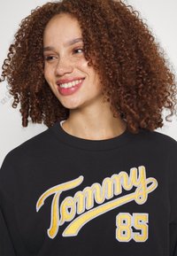 Svart sweatshirt med "Tommy" broderat i gult och vitt, med siffran "85" i en kontrasterande design.
