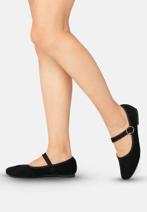 Legs wearing black Mary Jane flats with a single strap and gold buckle against a plain light background.