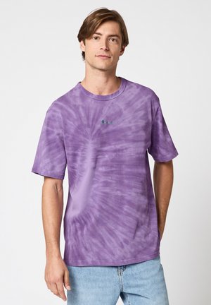 Jeune homme aux cheveux bruns portant un t-shirt violet teinté et un jean bleu clair, debout devant un fond blanc uni.