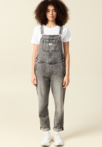 Pantalones de mezclilla negros con un ajuste relajado, tirantes ajustables, bolsillo frontal en el pecho y puños enrollados, combinados con una camiseta blanca y zapatillas blancas.