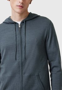 Felpa con zip di colore grigio scuro, dotata di cappuccio, tasche frontali e lacci. Presenta un logo discreto sul petto, con una texture del tessuto liscia.