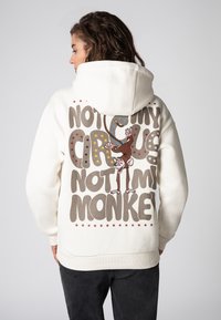 Crème hoodie met een grote grafische afbeelding op de achterkant, voorzien van speelse tekst en een illustratie van een aap, accentueert met gekleurde sterren.