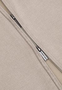 Beżowa dzianina z częściowo otwartym srebrnym zamkiem błyskawicznym, który odsłania białą metkę z czarnym tekstem wewnątrz ubrania.