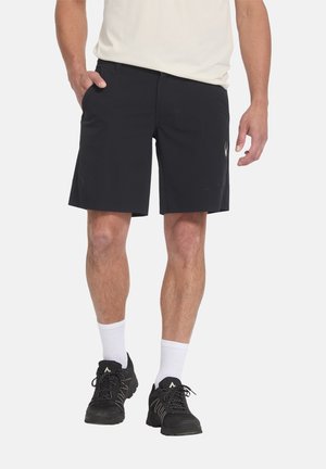 Mann trägt schwarze Shorts, weiße Sportsocken, schwarze Wanderschuhe und ein beiges kurzärmeliges Hemd, steht mit einer Hand in der Tasche.