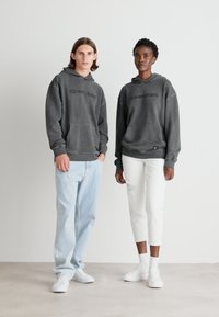 To personer har hettegensere med "TOMMYJEANS"-logo. Den ene hettegenseren er mørkegrå og overdimensjonert; den andre er kullfarget. Begge har på seg casual lyse jeans og hvite sneakers.