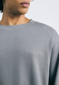 Szürke pamut pulóver kerek nyakkal és bordázott textúrával, finom hímzett Calvin Klein logóval a mellrészen.