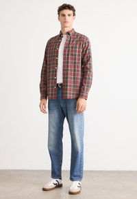 Geruite shirt in rood, groen en geel met lange mouwen, met knoopjeskraag; gecombineerd met lichtblauwe jeans en witte sneakers.
