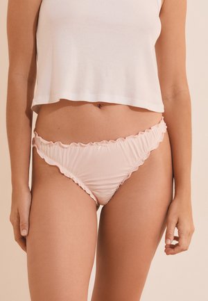 Femme portant un bas de bikini froncé rose clair et un débardeur court blanc sur un fond beige.