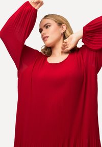 Rotes, plissiertes Kleid mit langen, gerafften Ärmeln und rundem Ausschnitt. Der Stoff weist vertikale Rippungen auf, die für Textur und Bewegung sorgen.