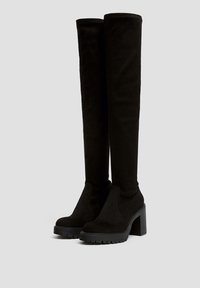 Botas negras de ante, hasta la rodilla, con tacón ancho y plataforma. Presentan una textura suave e inserciones laterales elásticas para facilitar su puesta.