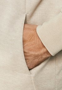 Tessuto felpa beige chiaro, con una tasca laterale e una mano all'interno, che mette in evidenza una consistenza morbida e un design rilassato.