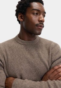 Brun ribbet sweater med rund hals, der viser et struktureret strikmønster. Modellens arme er korslagt foran, hvilket afslører tætsiddende ærmer.