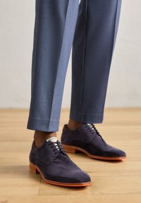 Scarpe in suede blu navy con punta arrotondata, dotate di suole e lacci arancioni in gomma. Abbinati a pantaloni su misura azzurri chiari, esposti su un pavimento di legno.