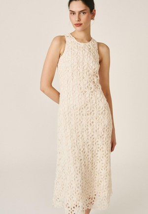 Mujer con vestido midi sin mangas de encaje color crema con patrón floral, de pie frente a un fondo neutro y claro.