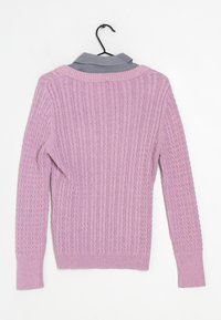 Pull en tricot rose clair avec texture côtelée et manches longues, avec une chemise grise à col visible sous l'encolure. Suspendu à un crochet.
