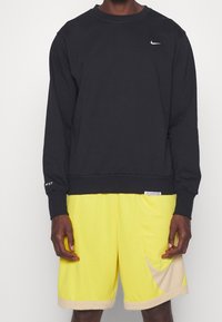 Sudadera negra con cuello redondo y mangas largas; presenta el logo de Nike. Pantalones cortos amarillos con un ribete beige y un patrón curvado en los laterales.