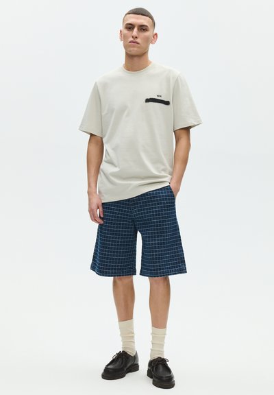 Young man standing, wearing beige oversized t-shirt with black abstract design, navy blue checkered shorts, cream socks, and black shoes.