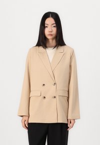 Beige kahe rinnaga bleiser, mis on valmistatud kergest kangast, kahte esimest taskut, suured nööbid ja lõikega krae, omades lõdvemat istuvust.