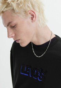 Felpa nera con logo ricamato "Levi's" in blu, collana in catena argentata e capelli biondi testurizzati. La vista profilo rivela i tratti del viso.