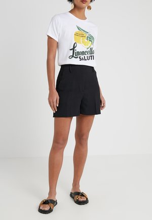 Kobieta ubrana w biały T-shirt z krótkim rękawem z grafiką cytryny i czarne szorty, do tego czarne sandały z ozdobnym złotym łańcuszkiem.