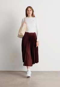 Femme marchant vêtue d'un haut à manches longues blanc, d'une jupe plissée bordeaux, de baskets blanches, et portant un sac fourre-tout beige à l'épaule.
