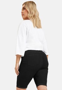 Pantalones cortos de mezclilla negros con dobladillos enrollados y bolsillos en la parte posterior, combinados con una camisa blanca de corte holgado, mostrados desde un ángulo lateral y trasero.