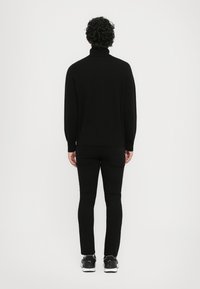 Μαύρο πουλόβερ με ψηλό λαιμό και ραμμένες μανσέτες και στρίφωμα, φοριέται πάνω από μαύρο στενό παντελόνι και μαύρα αθλητικά sneakers, προβαλλόμενο από πίσω.