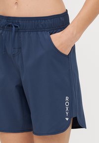 Tums zils peldbikses, kas izgatavotas no viegla auduma, ar elastīgu jostasvietu un auklu, kā arī sānu kabatām. "ROXY" logo baltā krāsā.