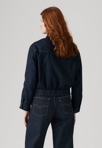 Veste en denim sombre avec une taille élastiquée, des poignets à bouton et des coutures contrastantes. Stylisée avec un jean sombre assorti et un design simple.