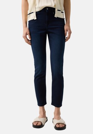 Persoon draagt donkerblauwe slim-fit jeans, een wit shirt, een lichtbeige vest en beige instapsandalen met zwarte zolen.