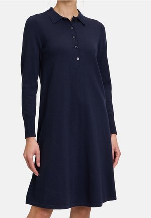 Femme portant une robe bleu marine à manches longues avec un col et une patte de boutonnage courte, dans un design uni, sur fond blanc.