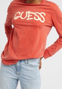 Rød langærmet skjorte med et slidt hvidt "GUESS" logo på forsiden; parret med lyseblå denim jeans. Blødt stof med en afslappet pasform.