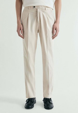 Hombre con pantalones beige de corte recto, camisa blanca por dentro y zapatos negros con cordones, de pie contra un fondo claro y liso.