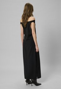 Robe maxi noire décolletée, avec une silhouette fluide, une fente latérale et une texture lisse, accompagnée de bottes à talons aiguilles noires.