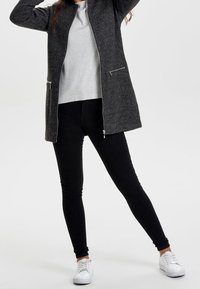 Langer grauer Mantel mit durchgehendem Reißverschluss, seitlichen Taschen, kombiniert mit einem hellgrauen Oberteil und schwarzen Skinny-Jeans. Weiße Sneakers vervollständigen das Outfit.