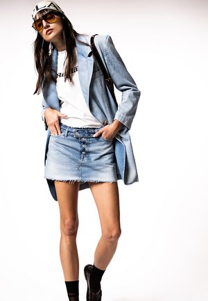 Kvinde iført en lys denimblazer, hvid grafisk t-shirt, denim mininederdel, sorte sokker, loafers, solbriller og et mærket hovedtørklæde.