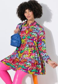 Buntes Kleid aus einem lebendigen, gemusterten Stoff mit verschiedenen Texten in Rosa-, Grün- und Blautönen. Eine blaue Stepptasche ergänzt das Outfit.
