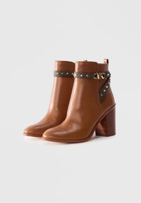 Brown leather ankle boots with block heels, decorated with dark studded straps and a gold "MK" logo buckle on each side.