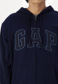 Sudadera con cremallera en azul marino que presenta la palabra "GAP" de manera prominente en letras azul claro, con un bolsillo tipo canguro y capucha con cordón.