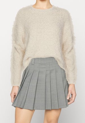 Žena nosící smetanový chlupatý svetr zastrčený do šedé plisované minisukně s vysokým pasem a károvaným vzorem.