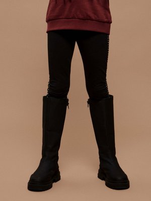 Person wearing black leggings and black knee-high boots with thick soles, dressed in a dark red top against a beige background.