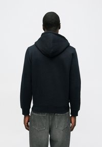 Tmavomodrá mikina s kapucňou s jednoduchým dizajnom, rebrovaným lemom a manžetami, nosená cez sivé džínsy. Pohľad zozadu odhaľuje bezšvovú kapucňu.