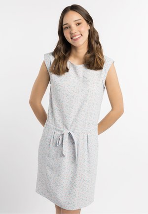 Jonge vrouw met lang bruin haar lacht, draagt een lichtblauwe mouwloze jurk met een kleine bloemenprint en een ceintuur die in de taille is gestrikt.