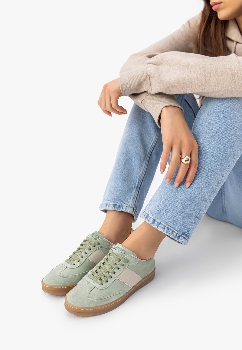 Personne portant des baskets en daim vert clair et un jean bleu clair, assise les bras reposant sur les genoux, montrant une bague en or à la main gauche.