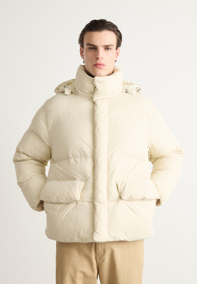 Cremefarvet pufferjakke med høj krave, store frontlommer og aftagelig hætte, der har en quilted struktur og glat stof.