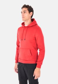 Giovane uomo rivolto a sinistra con un felpa rossa con cappuccio e tasca frontale, indossando pantaloni neri, su uno sfondo bianco semplice.