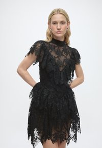 Γυναίκα με ξανθά μαλλιά που φορεί εφαρμοστό μαύρο δαντελένιο φόρεμα με βολάν και ψηλό λαιμό, όρθια με τα χέρια στη μέση σε λευκό φόντο.