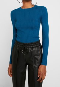 Haut à manches longues côtelé de couleur bleue vive, fabriqué en tissu doux, associé à un pantalon en similicuir noir avec taille élastique et cordon de serrage.